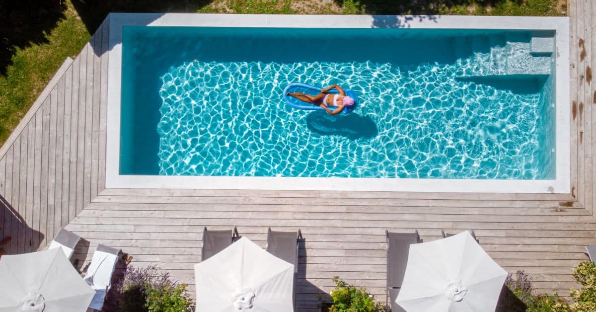 Aerial view of a modern swimming pool with a person relaxing on a float, surrounded by a wooden deck.