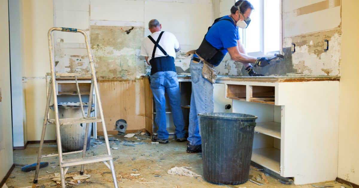 Two workers renovating a kitchen wall with tools and safety gear.