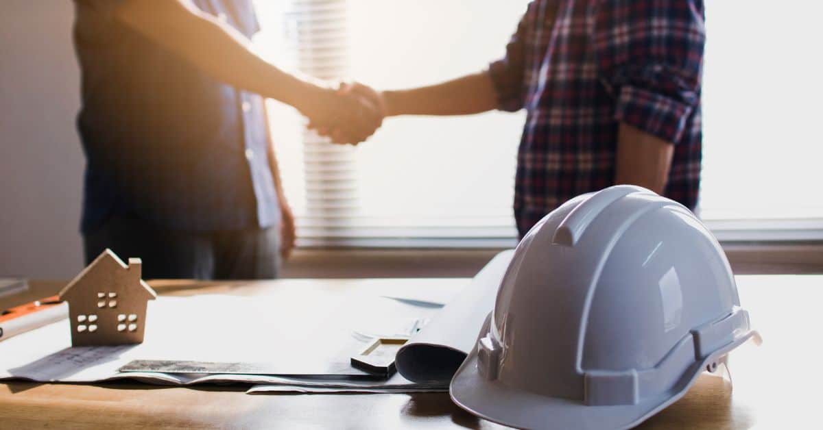 Two professionals shaking hands over a home building project with safety helmets on the table.